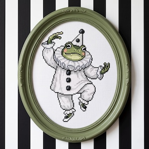 May include: A cross-stitch embroidery featuring a green frog dressed as a clown in a white ruffled collar, white tunic with black buttons, and a matching cone hat. The artwork is displayed within an oval, sage green decorative frame.