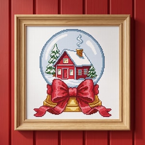 May include: A cross-stitch embroidery in a light wood frame depicting a snow globe with a red house, snow-covered roof, and chimney. Two green pine trees are inside the globe. A large red ribbon bow is tied around the base of the snow globe.
