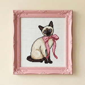 May include: Cross-stitch art of a Siamese cat with blue eyes and a pink bow, framed in a pink ornate frame. The cat is depicted in a seated position, with a cream-colored body and dark brown markings. The background is white.