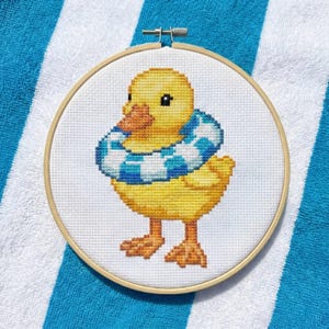 Op de afbeelding: Kruissteekborduurwerk van een gele eend met een blauw-witte geruite zwemband. De eend heeft een oranje snavel en poten. De borduursel zit in een houten hoepel en ligt op een blauw-wit gestreepte handdoek.
