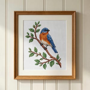 May include: Cross-stitch artwork of a bluebird perched on a branch with green leaves. The bird has blue and orange plumage. The artwork is framed in a light brown wooden frame with a white mat.