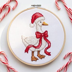 May include: A cross-stitch embroidery piece in a round wooden hoop featuring a white goose wearing a red and white Santa hat and a large red ribbon bow around its neck. The goose has orange feet and a yellow beak, set against white fabric.