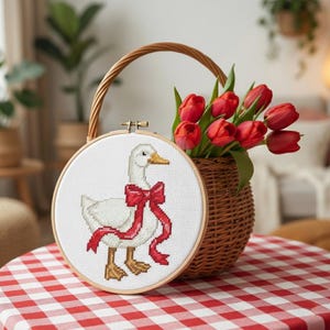 May include: A cross-stitch embroidery hoop featuring a white goose with a red bow. The hoop is displayed on a red and white checkered tablecloth next to a wicker basket filled with red tulips. The basket has a handle.