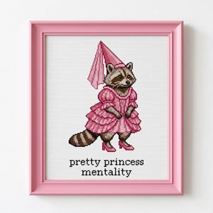 Peut inclure: Point de croix d'un raton laveur en robe et chapeau de princesse roses, portant des talons roses. L'œuvre encadrée porte le texte "pretty princess mentality" en noir.