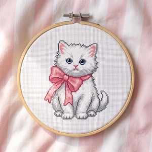Op de afbeelding: Een kruissteek borduurwerk met een wit kitten met blauwe ogen en een grote roze strik om de nek. Het ontwerp is geborduurd op witte Aida-stof en wordt stevig vastgehouden in een ronde houten borduurring met een metalen schroef.