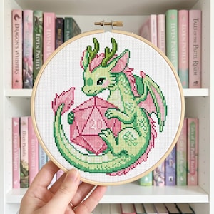 Pode incluir: Um bastidor de bordado em ponto cruz com um dragão verde e rosa segurando um dado rosa de vinte lados. O dragão tem escamas verdes, asas rosa e barriga rosa. O bordado está em um bastidor de madeira. O fundo mostra uma estante com livros rosa e verdes.