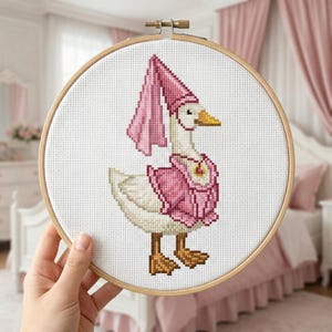 May include: A cross-stitch embroidery of a goose wearing a pink outfit and hat, set in a wooden hoop. The goose has a yellow beak and brown feet. The background is white, and the embroidery is held by a hand.