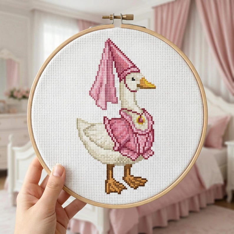 May include: A cross-stitch embroidery of a goose wearing a pink outfit and hat, set in a wooden hoop. The goose has a yellow beak and brown feet. The background is white, and the embroidery is held by a hand.