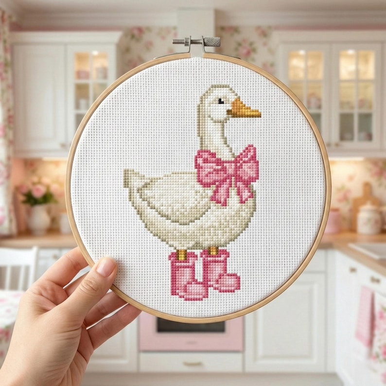 May include: A cross-stitch embroidery hoop featuring a white goose with a pink bow and pink boots. The goose has a yellow beak and is set against a white background. The embroidery hoop is held by a hand.