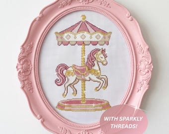 May include: A cross-stitch embroidery of a carousel horse in pink, white, and gold, framed in an oval pink ornate frame. The design features a carousel horse with a pink mane and tail, a gold pole, and a pink canopy. Text reads: WITH SPARKLY THREADS!