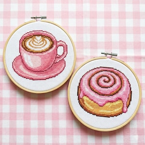 Puede incluir: Dos aros de punto de cruz muestran una taza de café rosa con un diseño en forma de corazón en la espuma y un rollo de canela glaseado rosa. El bordado está sobre un fondo blanco con un patrón de cuadros rosas.