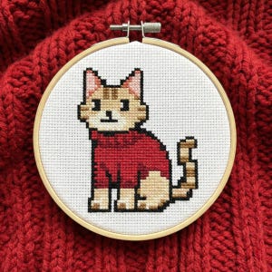 Puede incluir: Bordado de punto de cruz de un gato con un suéter rojo, en un aro de madera. El gato es marrón y beige, con un suéter rojo. El fondo es una tela roja texturizada.