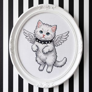 Peut inclure: Une broderie au point de croix représentant un chaton blanc duveteux aux yeux bleus avec des ailes à plumes, portant un collier noir à pointes. L'œuvre est présentée dans un cadre ovale blanc avec des détails en relief sur le bord.