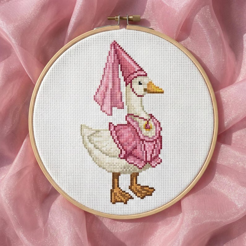 May include: A cross-stitch embroidery of a goose wearing a pink outfit and hat, set in a wooden hoop. The goose has a yellow beak and brown feet. The pink outfit includes a cape and a decorative collar. The background is a pink fabric.
