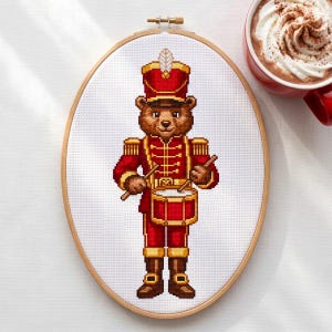 May include: An oval wooden embroidery hoop containing a cross-stitch pattern of a brown bear dressed in a red and gold military-style marching band uniform with a tall hat, playing a red drum with two drumsticks against a white fabric background.