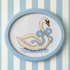 May include: Cross-stitch artwork of a swan with a blue bow, framed in a light blue oval. The swan is white and beige, with an orange beak and black eyes. The background is white with blue stripes.