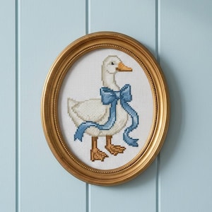 May include: A framed cross-stitch of a white goose wearing a blue bow. The goose has a yellow beak and feet. The oval frame is gold and hangs on a light blue wall. The artwork is a charming piece of home decor.
