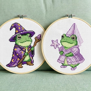 Może przedstawiać: Dwa tamborki do haftu krzyżykowego przedstawiające zielone żaby przebrane za czarodzieja i wróżkę. Żaba-czarodziej ma fioletowy kapelusz i pelerynę ze złotymi gwiazdami, trzymając laskę. Żaba-wróżka ma fioletową sukienkę i trzyma różdżkę w kształcie gwiazdy.