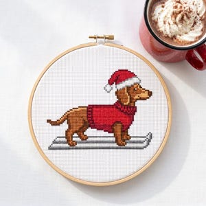 May include: A cross-stitch embroidery in a round wooden hoop featuring a brown dachshund wearing a red jumper and a red and white Santa hat, standing on grey skis. Beside the hoop sits a red mug filled with a beverage topped with whipped cream.