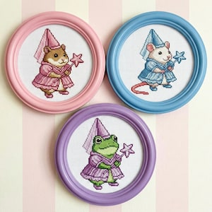 May include: Three framed cross-stitch artworks featuring whimsical animal characters. A hamster, a mouse, and a frog are dressed as fairytale figures, each holding a star-shaped wand. The frames are pastel pink, blue, and purple.