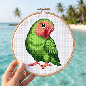 May include: A cross-stitch embroidery of a green and pink parrot in a wooden hoop. The parrot has a red beak and is set against a white background. The background is a tropical beach scene with palm trees and blue water.