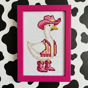 May include: Cross-stitch artwork of a white goose wearing a pink cowboy hat, vest, and boots. The framed art has a bright pink border. The background is white cross-stitch fabric. The goose has a yellow beak and is set against a black and white cow print background.