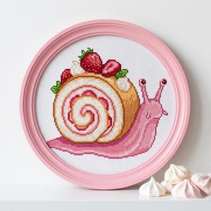 May include: Cross-stitch artwork of a pink snail with a strawberry Swiss roll shell, framed in a pink circular frame. The cake is topped with strawberries and cream. Meringues are in the foreground.