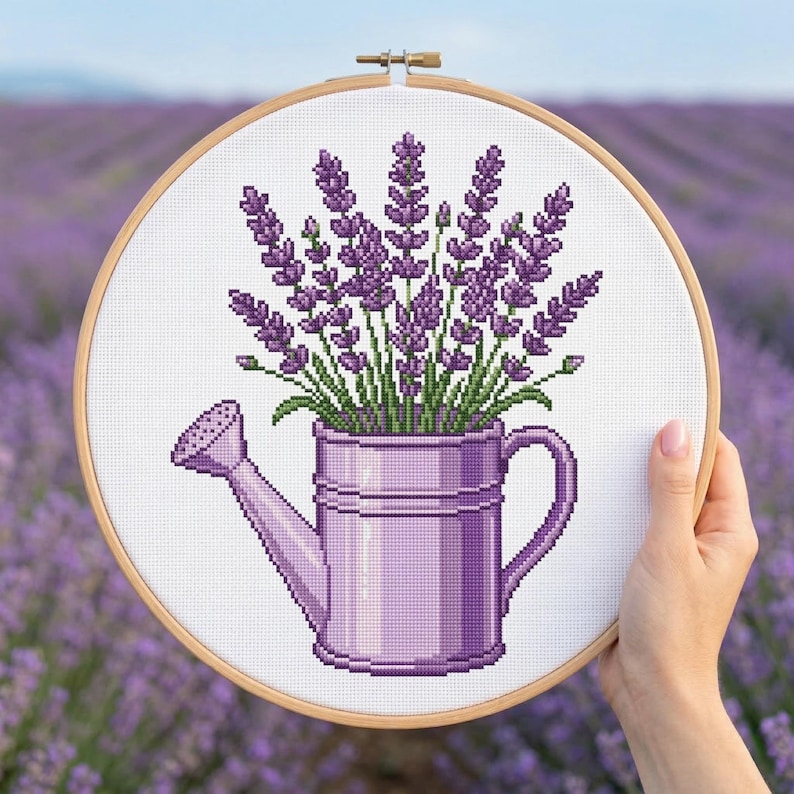 May include: A cross-stitch embroidery hoop showing a purple watering can filled with lavender. The lavender has green stems and purple flowers. The hoop is held in front of a lavender field.