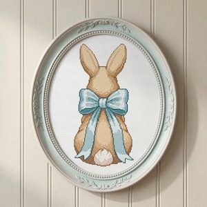 Puede incluir: Un punto de cruz enmarcado de un conejito visto por detrás, con un lazo azul claro. El conejito es de color canela con una cola blanca, sobre un fondo blanco. El marco ovalado es azul claro y tiene un borde decorativo.