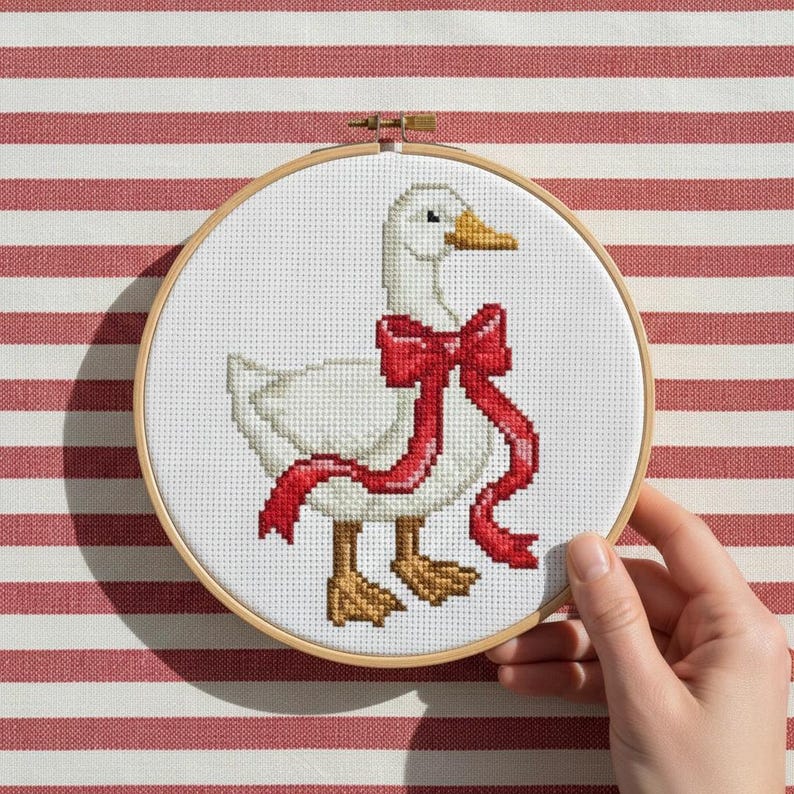 May include: Cross-stitch embroidery of a white goose wearing a red bow. The goose has a yellow beak and brown feet. The embroidery is set in a wooden hoop against a red and white striped background.