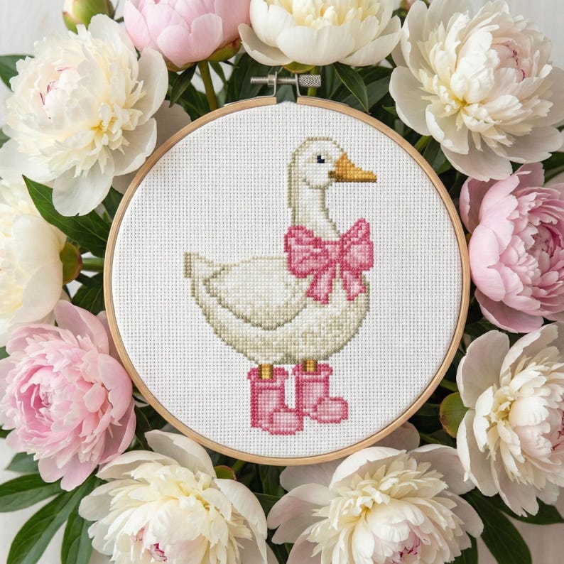 May include: A cross-stitch embroidery hoop featuring a white goose with a pink bow and boots. The goose is surrounded by a wreath of white and pink peonies, creating a charming, handcrafted aesthetic.