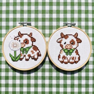 May include: Two cross-stitch embroidery hoops featuring a cartoon cow design. The cows are white with brown spots and wear green bows. One cow holds a dandelion. The hoops are set against a green and white checkered background.