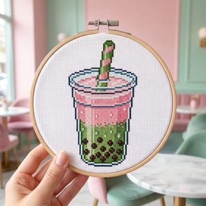 Może przedstawiać: Haft krzyżykowy przedstawiający napój bubble tea w przezroczystym kubku, trzymany w drewnianej tamborku. Napój jest różowy i zielony z paskowaną słomką. Wzór jest pikselowany.