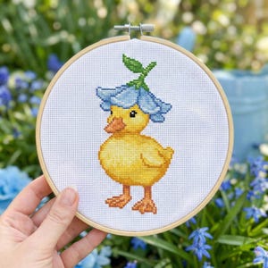 Puede incluir: Un bordado de punto de cruz de un patito amarillo con un sombrero de flores azules, en un aro de madera. El patito tiene pico y patas naranjas. El fondo es una escena borrosa al aire libre con vegetación y flores azules.