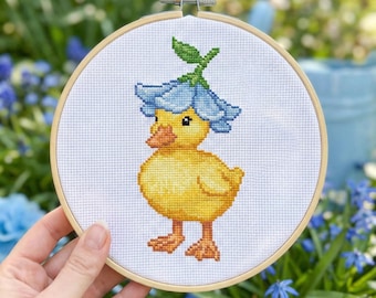 May include: A cross-stitch embroidery of a yellow duckling wearing a blue flower hat, set in a wooden hoop. The duckling has an orange beak and feet. The background is a blurred outdoor scene with greenery and blue flowers.