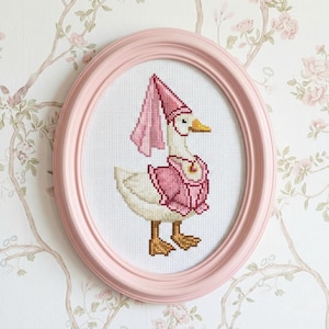 May include: A cross-stitch artwork of a goose wearing a pink hat and vest, framed in a pink oval frame. The goose has a yellow beak and brown feet. The background is a floral wallpaper.