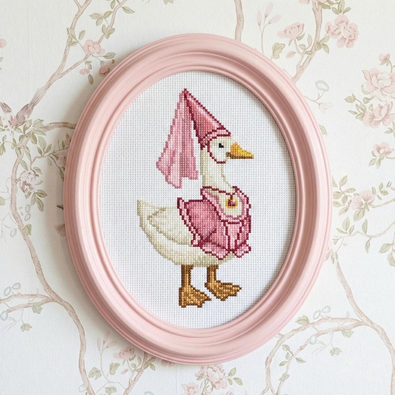 May include: A cross-stitch artwork of a goose wearing a pink hat and vest, framed in a pink oval frame. The goose has a yellow beak and brown feet. The background is a floral wallpaper.