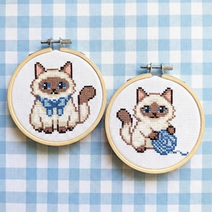 Puede incluir: Dos aros de punto de cruz con gatos siameses. Un gato lleva un lazo azul, el otro sostiene una bola de lana azul. Los gatos tienen ojos azules, orejas y colas marrones. Fondo a cuadros azul y blanco.