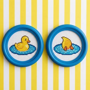 Puede incluir: Dos obras de arte en punto de cruz enmarcadas. Cada una presenta un pato amarillo en una escena de agua azul. Los patos están en diferentes posiciones, uno flotando y otro buceando. Los marcos son redondos y azules, sobre un fondo a rayas amarillas y blancas.