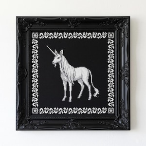 May include: A framed cross-stitch of a unicorn on a black background. The white unicorn has a long mane and tail, and a single horn. The frame is black with ornate detailing and a white floral border.