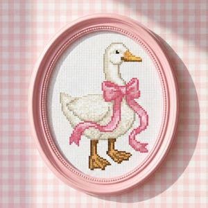 Könnte beinhalten: Ein Kreuzstich in einem rosa ovalen Rahmen. Das Kunstwerk zeigt eine weiße Gans mit einer rosa Schleife. Die Gans hat einen gelben Schnabel und braune Füße. Der Hintergrund ist ein weißes Gitter.
