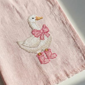 May include: A pink, fringed towel featuring a cross-stitched goose wearing a pink bow and pink boots. The goose has a white body, a yellow beak, and black eyes. The towel has a light pink background.