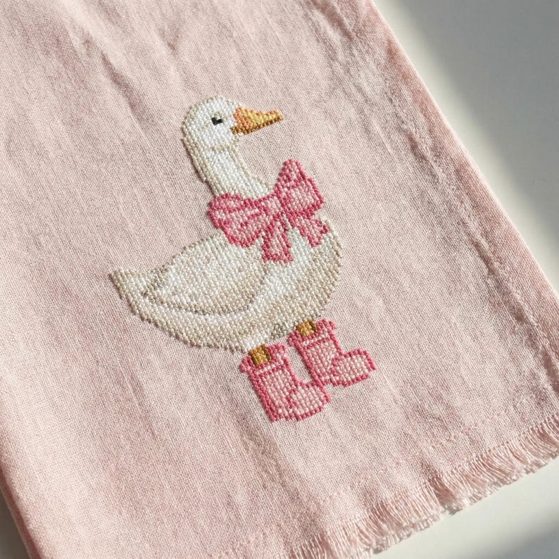 May include: A pink, fringed towel featuring a cross-stitched goose wearing a pink bow and pink boots. The goose has a white body, a yellow beak, and black eyes. The towel has a light pink background.