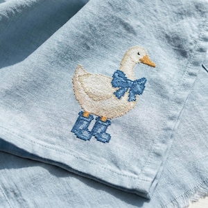 May include: A light blue linen fabric with a cross-stitched goose design. The goose is white with a yellow beak, wearing a blue bow and blue boots. The fabric has a frayed edge.