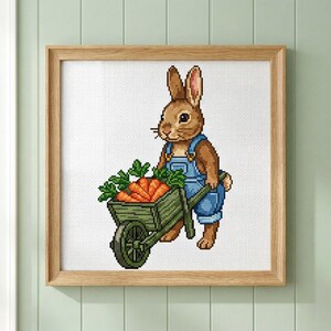 May include: Cross-stitch artwork of a brown bunny wearing blue overalls, pushing a green wheelbarrow filled with orange carrots. The artwork is framed in a light brown wooden frame and set against a white background.