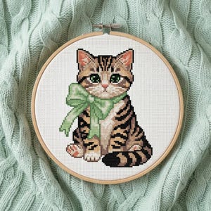 Può includere: Ricamo a punto croce di un gatto tigrato con un fiocco verde. Il gatto ha strisce marroni e nere, occhi verdi e orecchie rosa. Il ricamo è in un telaio di legno.