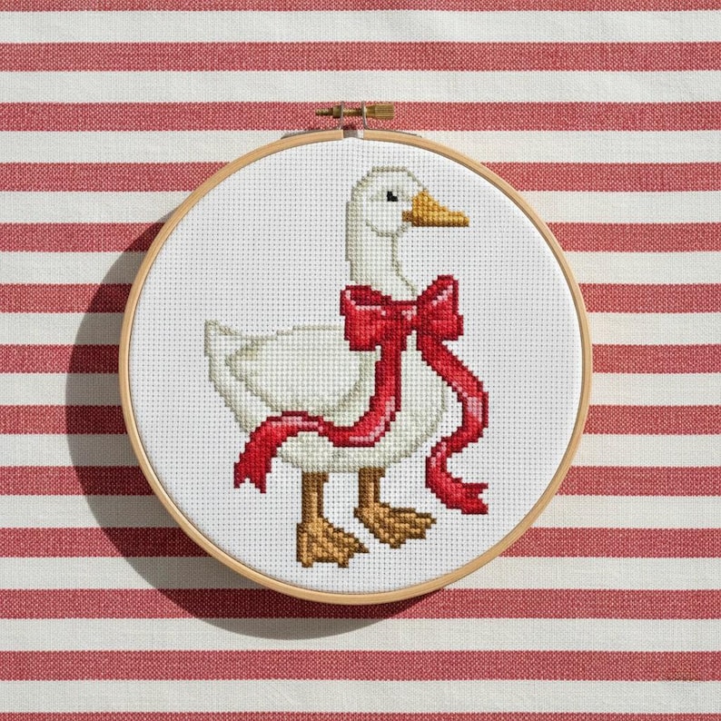 May include: A cross-stitch embroidery of a white goose wearing a red bow, set in a wooden hoop. The goose has brown feet and a yellow beak. The background is a white fabric with red stripes.
