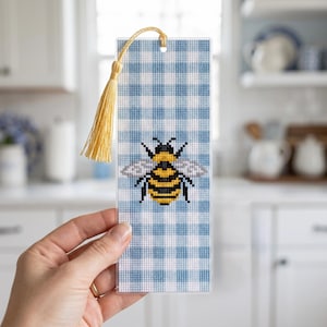 May include: A cross-stitched bookmark with a bee motif. The bookmark has a blue and white gingham pattern and a yellow tassel. The bee is black and yellow with white wings. The bookmark is held by a person.