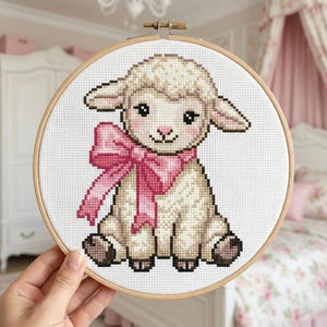 May include: A cross-stitch embroidery of a lamb wearing a pink bow, set in a wooden hoop. The lamb has a fluffy, off-white coat, dark eyes, and a pink nose. The background is white, and the overall design is soft and charming.