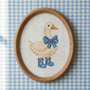 May include: Oval framed cross-stitch art featuring a goose wearing a blue bow and blue boots. The goose is beige with an orange beak. The frame is made of wood. The background is white fabric.